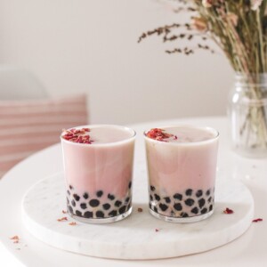 two glasses filled with strawberry rose milk tea and tapioca pearls next to a vase of flowers
