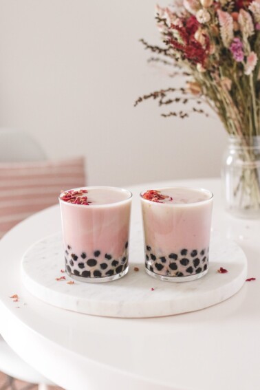 two glasses filled with strawberry rose milk tea and tapioca pearls next to a vase of flowers