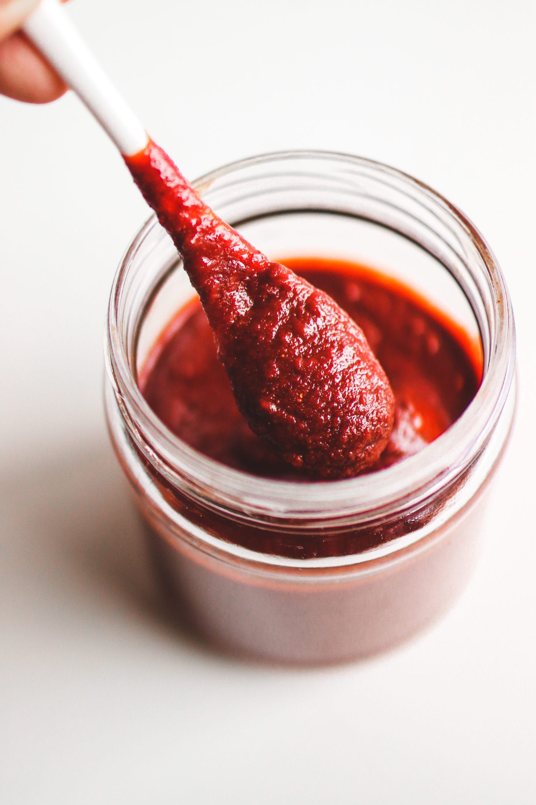 a closeup of a spoon holding red pepper sauce over a jar of sauce.