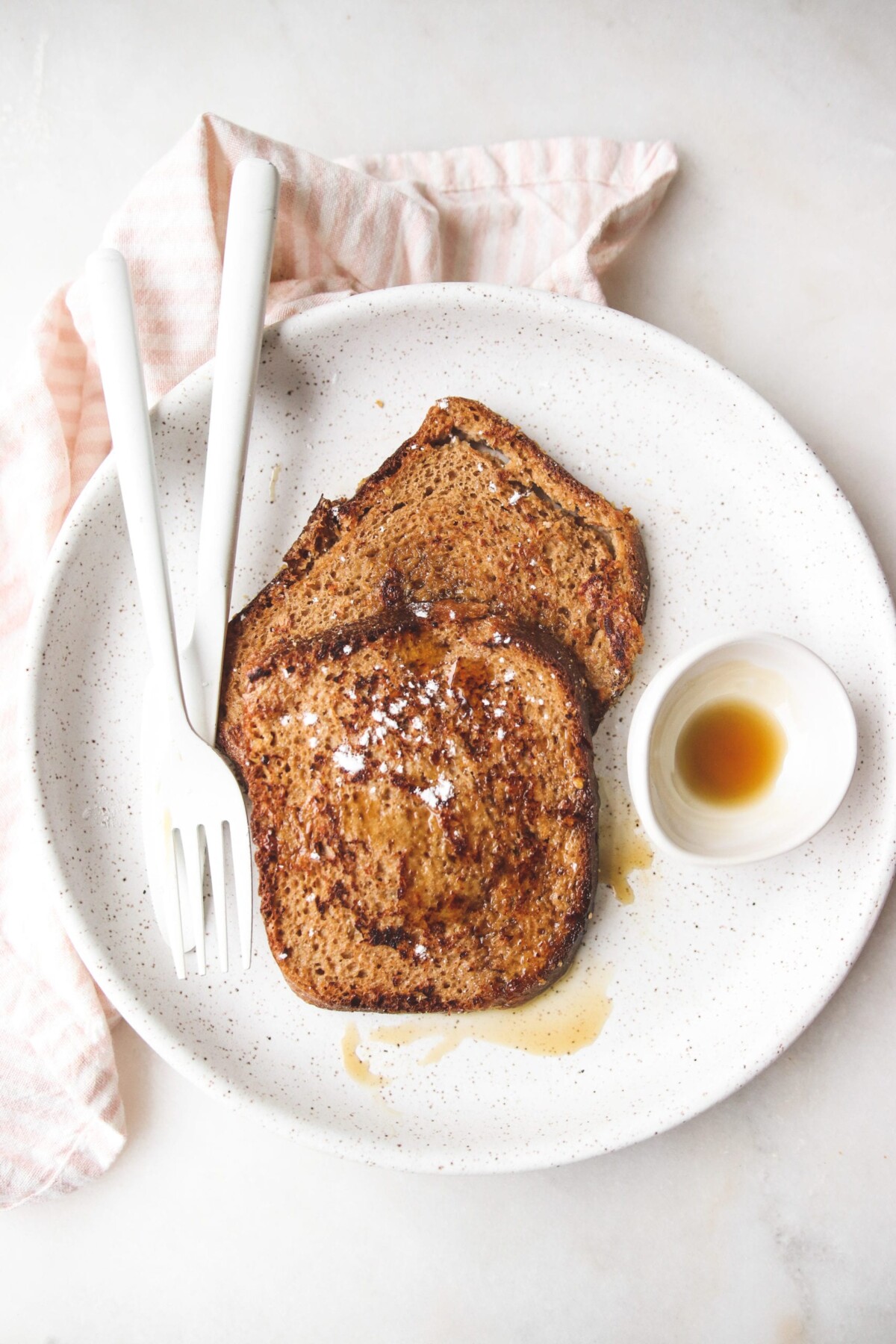 an overhead shot of vegan french toast on a white plate with two forks and a bowl of maple syrup. 
