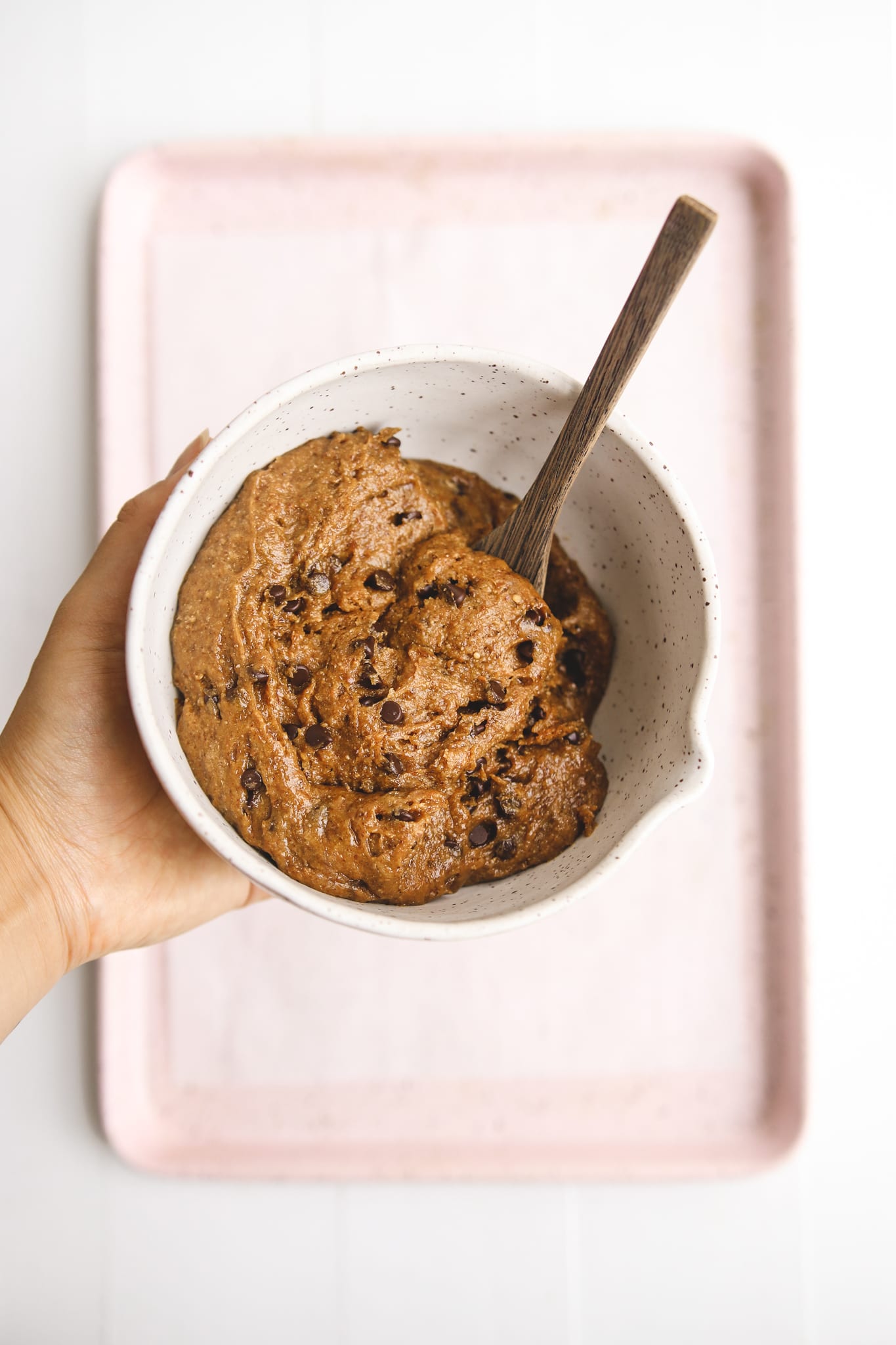 a hand holding a mixing bowl filled with vegan cookie dough and a wooden spoon.