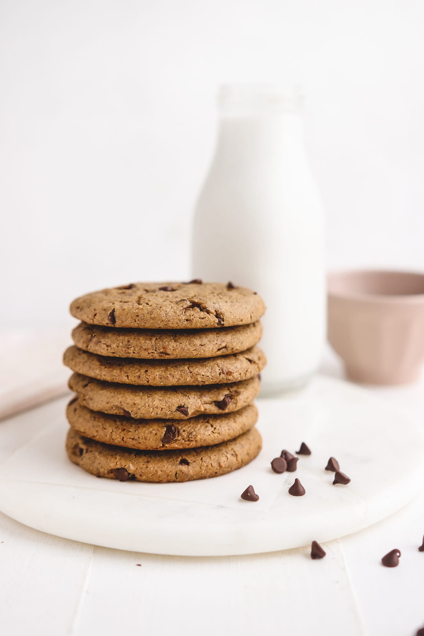 an up close photo of a stack of chocolate chip cookies with a glass jug of milk and pink bowl behind it. 
