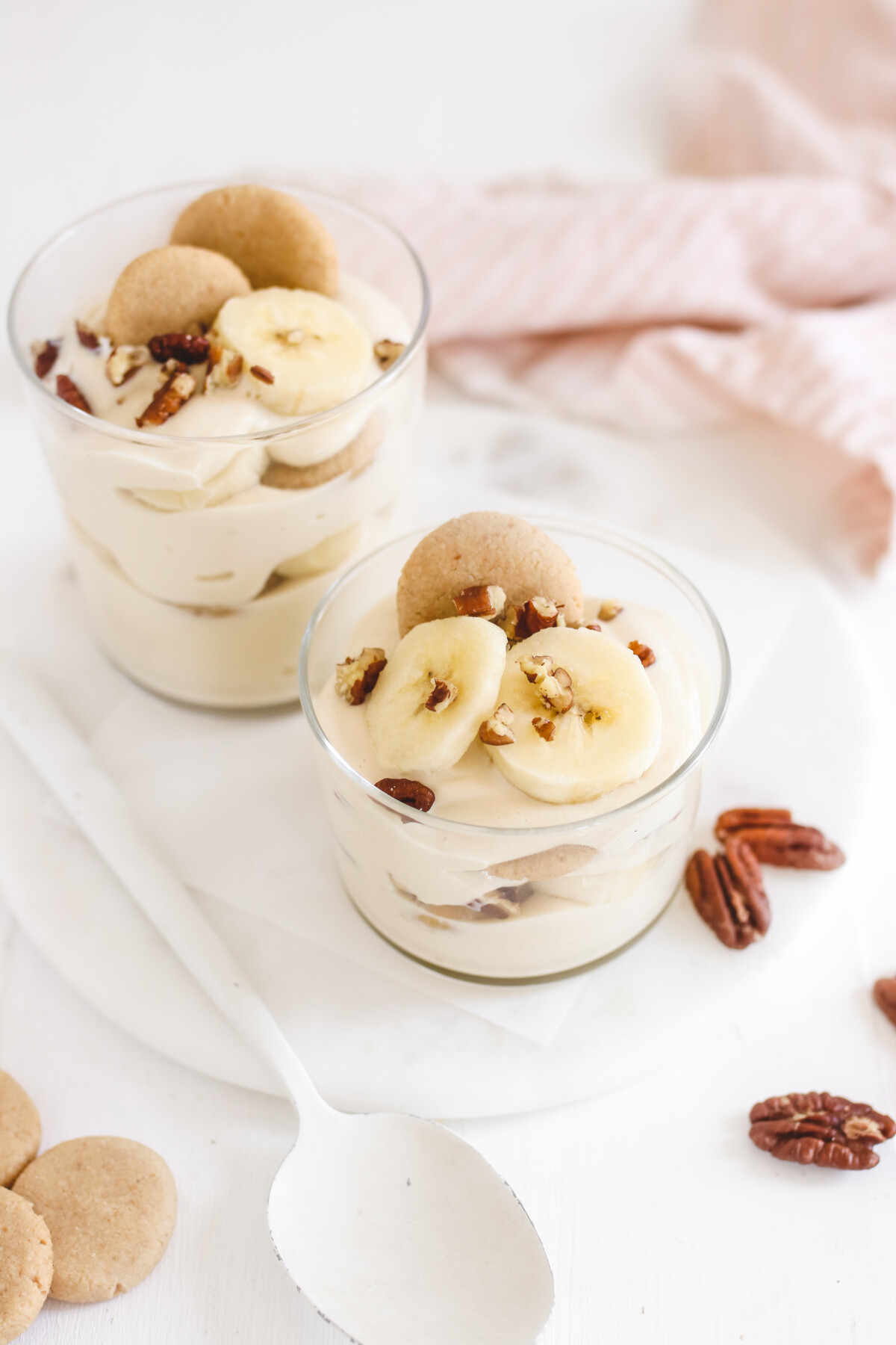 two glasses filled with vegan banana pudding and topped with vanilla wafer cookies, banana slices, and chopped pecans
