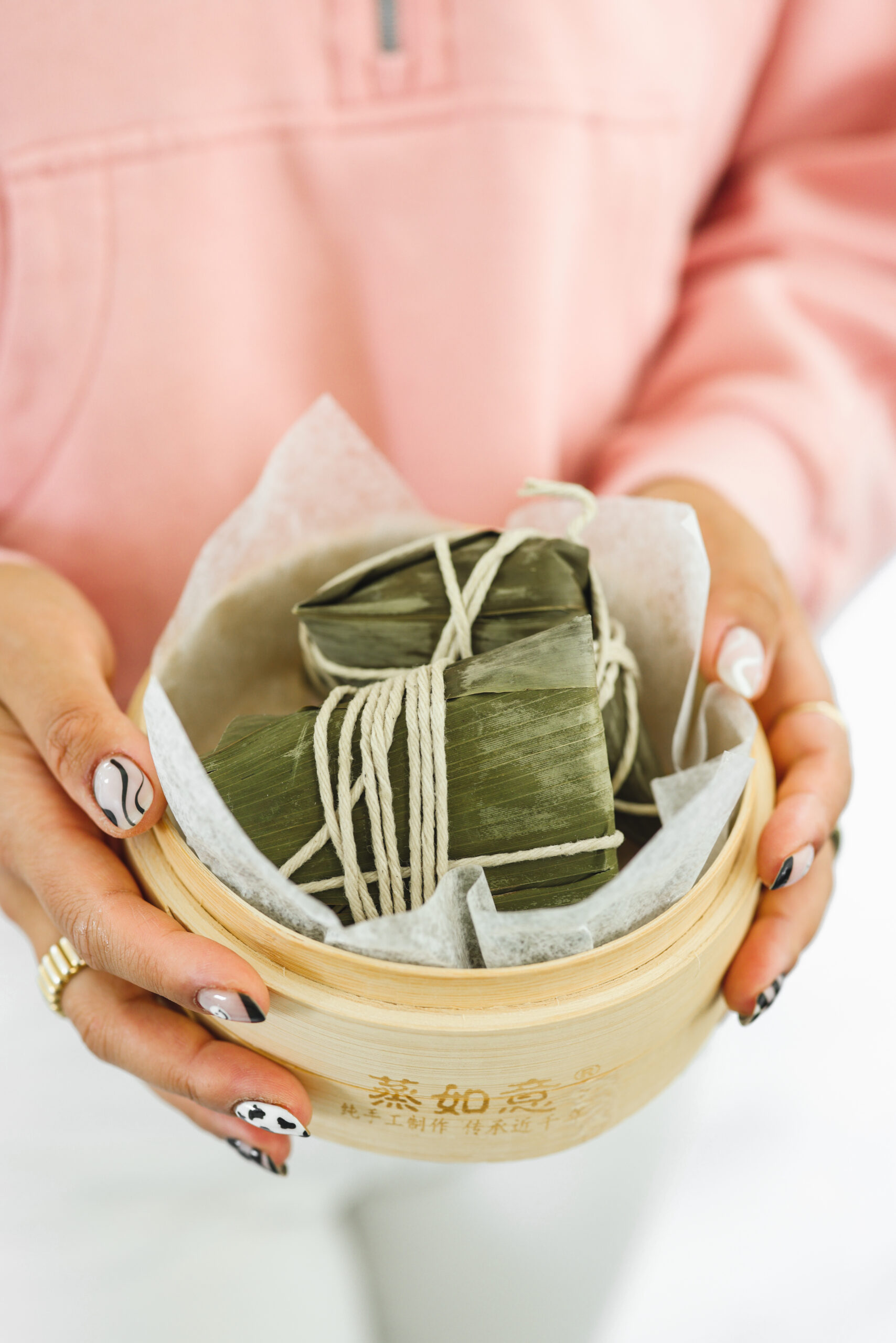 two hands holding a bowl filled with sticky rice dumplings tied in bamboo leaves. 